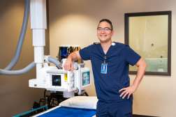 Ryan Whitcraft standing in a radiology room while wearing scrubs, with one arm propped up on radiology equipment that hangs from the ceiling. 