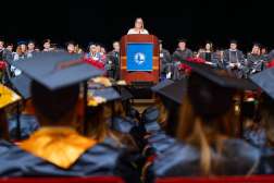 Crowd of NTC graduates seated wearing black mortarboards with gold accents, viewed from behind toward a podium and stage.