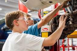Two students wearing safety glasses work together on a car’s suspension system in an automotive workshop.