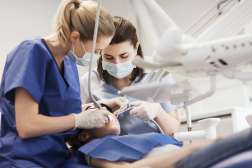 Two dental professionals wearing scrubs, gloves, and face masks performing a dental procedure on a patient lying in a dentist’s chair, with dental instruments and overhead equipment visible.