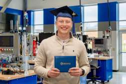 Ethan Sondelski smiling while wearing his graduation cap and holding his diploma while standing inside the Industry 4.0 Smart Manufacturing Lab
