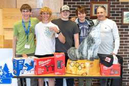 Students and instructor pose with tools and Olympic-themed wolf sculpture at technical college workshop.