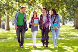 Four college students walk together outside on a sunny day, smiling and holding books on a green, tree-lined campus.