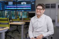 Jordan smiles, while seated in a video production room, with multiple computer monitors behind him displaying video editing software and nature scenes.