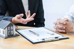 A real estate agent in a dark suit explains details to a client, with a miniature house, papers, pen, and key on the desk.