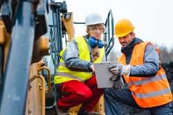 Two construction workers, a woman and a man, in hard hats and safety vests, review a clipboard together near heavy machinery.