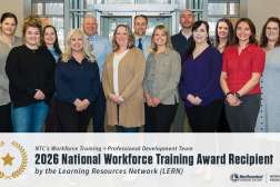 Fifteen smiling adults in business-casual attire stand in two rows in a conference room, posing as an award-recipient group.