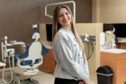 A smiling student stands in a dental lab setting, with a dental chair and equipment visible behind her, wearing a casual sweatshirt and looking toward the camera.