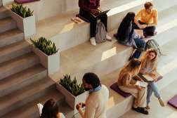 Students seated on wide tiered concrete steps in a sunlit campus atrium, reading, using laptops and chatting.