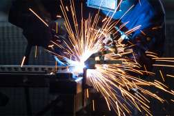 Welder in blue protective jacket and gloves using a welding torch on metal, bright arc and orange sparks shooting out.