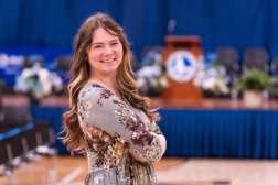 A young woman (Ashley Aull) with long wavy hair smiles while standing with her arms crossed in front of a graduation ceremony stage decorated with flowers and a podium. She wears a floral dress and poses in a gymnasium setting prepared for commencement activities.