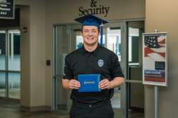 Kyle smiling while wearing his graduation cap and holding up his diploma in front of the Safety and Security Office.