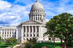 External view of the capitol building in Madison Wisconsin with grand white columns, manicured green lawn and trees.