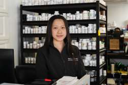 Nancy Xiong, a pharmacy technician youth apprentice, stands behind a pharmacy counter, looking toward the camera, with shelves of prescription medication bottles neatly organized behind her. She wears a black pharmacy-branded jacket and is positioned in a working pharmacy environment that includes medication storage, dispensing supplies, and computer workstations.