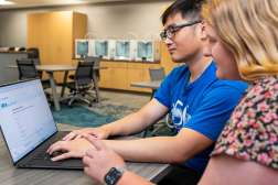 A student and faculty member collaborate at a laptop in a bright computer lab; a young man types while a young woman watches.