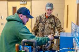 Two men focused on metal machinery in a workshop: one in a green hoodie adjusting equipment, another in camouflage jacket.