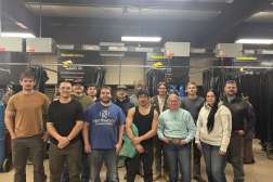 A group of students and an instructor pose together. The students stand in front of a row of welding booths equipped with ventilation systems and welding curtains, representing a variety of technical training backgrounds. Industrial equipment, welding stations, and safety infrastructure fill the workspace behind them.