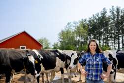 A photo shows a person standing with hands on their hips beside a group of black-and-white cows. A red barn and a line of trees are visible in the background.