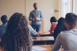College students seated at desks facing a standing instructor at the front of a sunlit classroom.