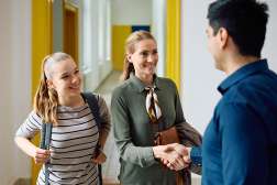 Smiling female teacher in green blazer shakes hands with a man in a school hallway with a smiling girl with a backpack.