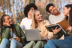 Five diverse college students sit on grass in an autumn park, laughing and studying together with notebooks and a laptop.