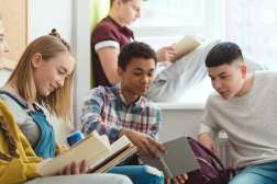 Five diverse teenagers in a bright classroom, casually seated by a window, smiling while reading open textbooks.
