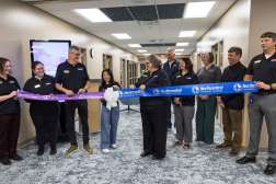 Group of people in an office hallway cutting a long blue ceremonial ribbon with oversized scissors.