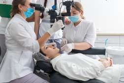 Patient reclined in dental chair receiving care from two masked clinicians in white coats.