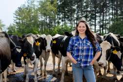 Mary stands with her hands on her hips in front of dairy cows with pine trees in background.