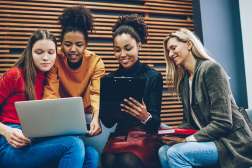 Four casually dressed young women of diverse backgrounds sitting together on a bench indoors, smiling and sharing a laptop.