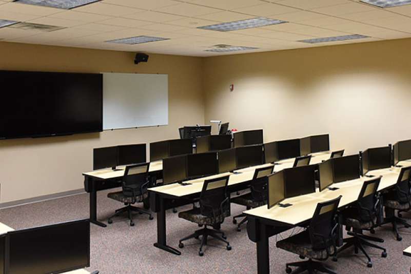 A computer lab classroom setup with several rows of tables with computers, along with an instructor's stand and a TV mounted to the wall.