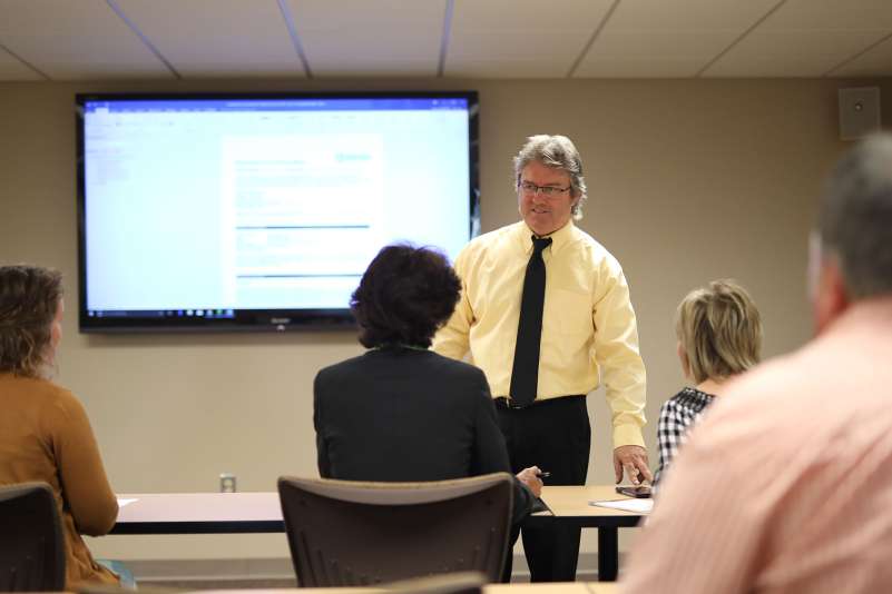 A businessman is standing up in front of a room full of businesspeople giving a lecture