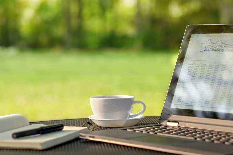 A close up view of a laptop, notebook, and a cup of coffee on a table outside near grass and trees