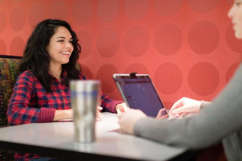 A student sits in a booth near the Campus Cafe, across from another individual, who is in the foreground, just off camera.