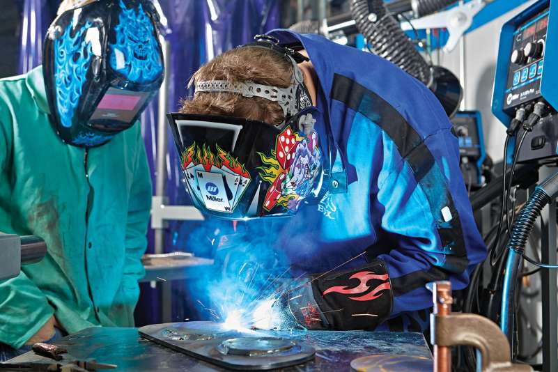 A student is welding medal together as an intern watches. Bright sparks shoot out from the objects being welded together