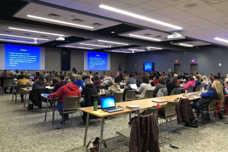 View from the back of the professional conference center with a speaker addressing conference attendees at the front of the room.