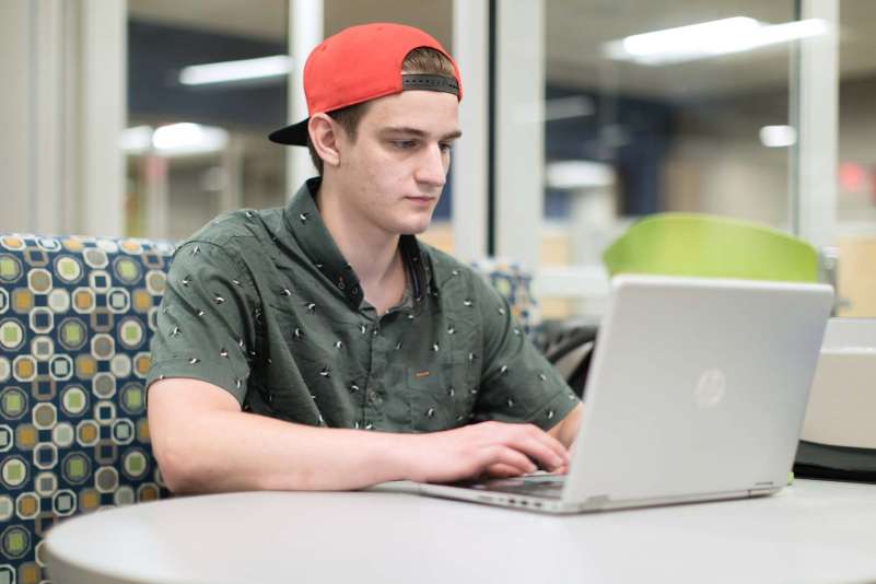 Student sitting in a booth, typing on his laptop, which sits on the table in front of him.