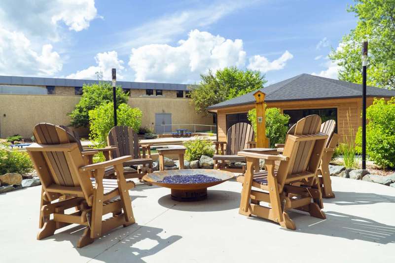 Six rocking chairs surround a large fire pit in the courtyard. Rocks and a landscaped garden surround the paved area. A gazebo and the main NTC Wausau Campus building are visible in the background.