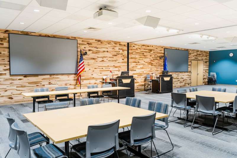 The front wall of the Community Training Room features podiums, large projection screens, and a wood paneled wall. Several tables and chairs are available to seat large audiences.