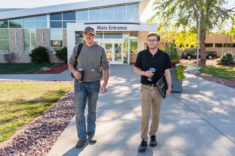 Two students sharing a laugh while carrying coffee mugs and school supplies as they walk outside the main entrance of the Wausau Campus.