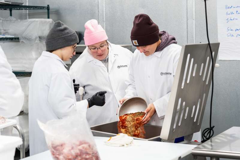 Three food industry workers in white coats and colorful beanies pour spice mixture into a metal container.