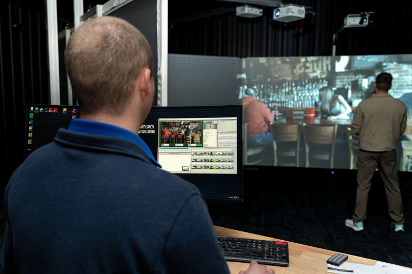 A simulation technician monitors a computer interface while another person engages in a bar scenario projected on a large screen at the Community Safety Simulation Center.