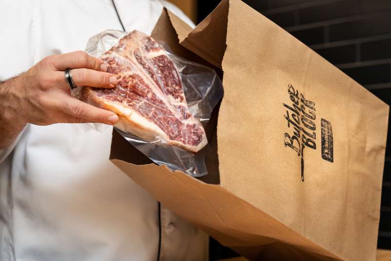 Butcher Block staff’s hand placing a vacuum-sealed T-bone steak into a brown paper bag displaying "The Butcher's Block" logo.