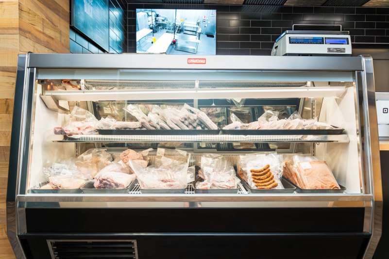 Butcher Block refrigerated display case filled with packaged meats on two shelves. A screen and a scale are visible above.