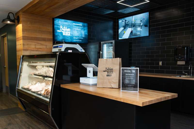 Butcher Block storefront, including shop counter with a glass display case of meats, scale, and tablet. A branded paper bag sits under two wall-mounted screens.