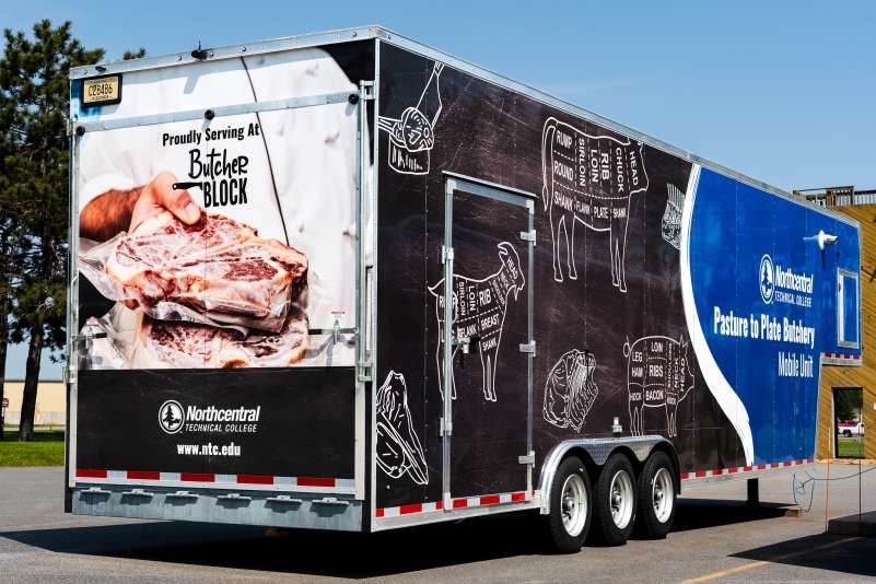 The Pasture to Plate mobile lab trailer, as seen from a rear angle view. The rear displays the Butcher Block and NTC logos, along with photos of steak. Diagrams of butchery cuts adorn the side of the trailer.