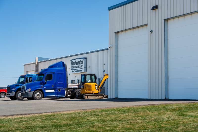 NTC's Diesel Technology Center building, with two blue semi trucks and a yellow excavator parked outside.