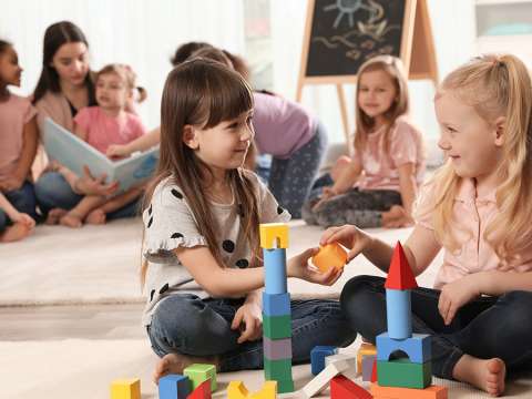 Children are playing with toy blocks and having a story read to them by a child care worker