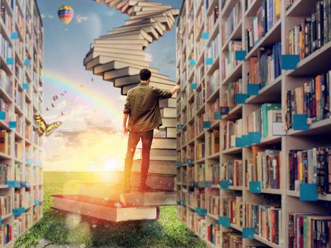 Inside of a library, a student is walking up a staircase of books leading up into the sky.