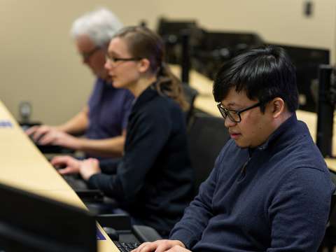 Three students in a classroom are typing on keyboards at desktop workstations.